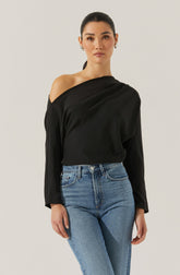 A woman is wearing a black satin blouse with a one-shoulder design, featuring long sleeves and a relaxed fit. She has paired it with high-waisted, light-wash blue jeans.